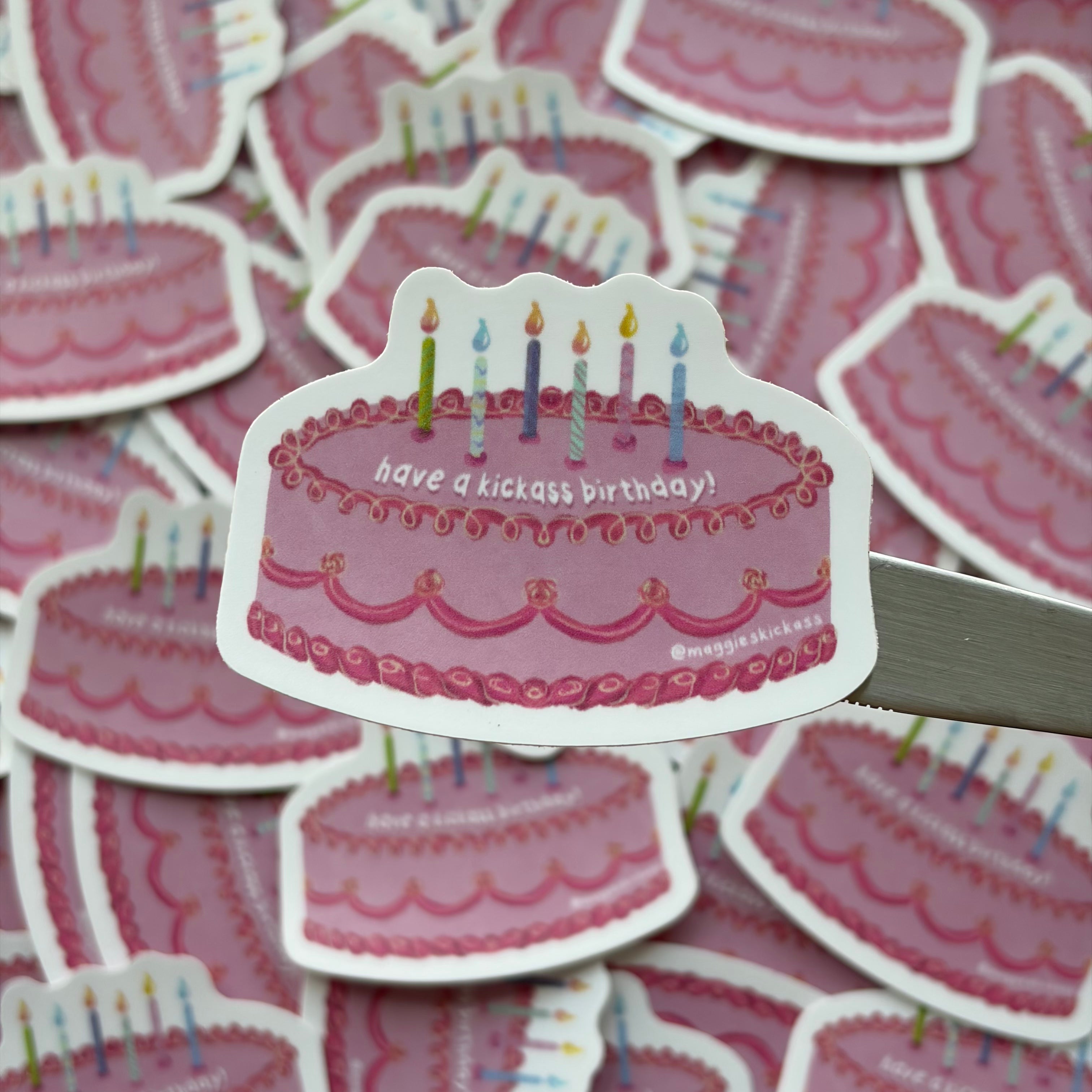 A die-cut sticker of a pink birthday cake with colorful candles is held by a metal butter knife over a pile of identical stickers. The sticker reads 'have a kickass birthday!' and features a small handle credit '@maggieskickass'.