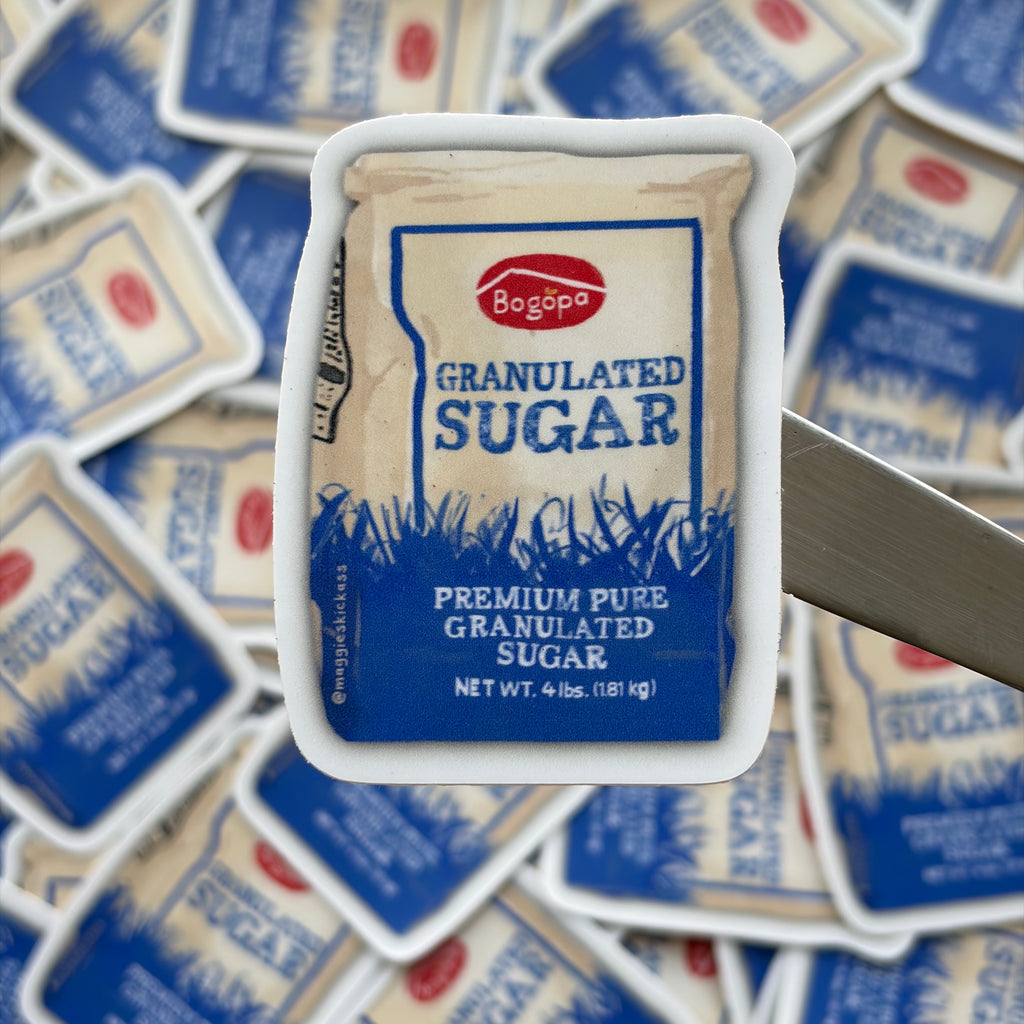 A close-up of a sticker featuring a white paper bag of Bogopa granulated sugar, set against a blurred background of scattered sugar bags stickers.