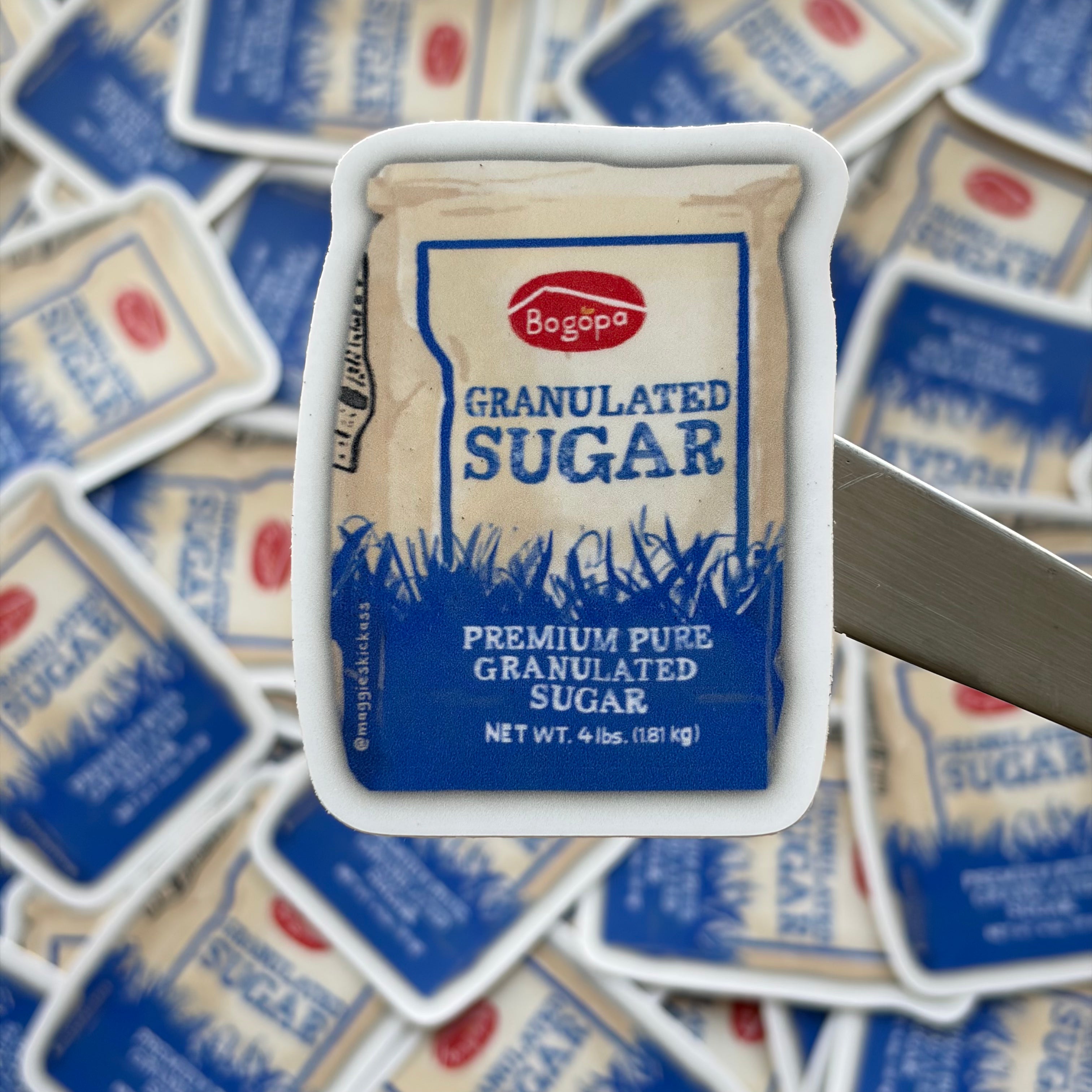 A close-up of a sticker featuring a white paper bag of Bogopa granulated sugar, set against a blurred background of scattered sugar bags stickers.