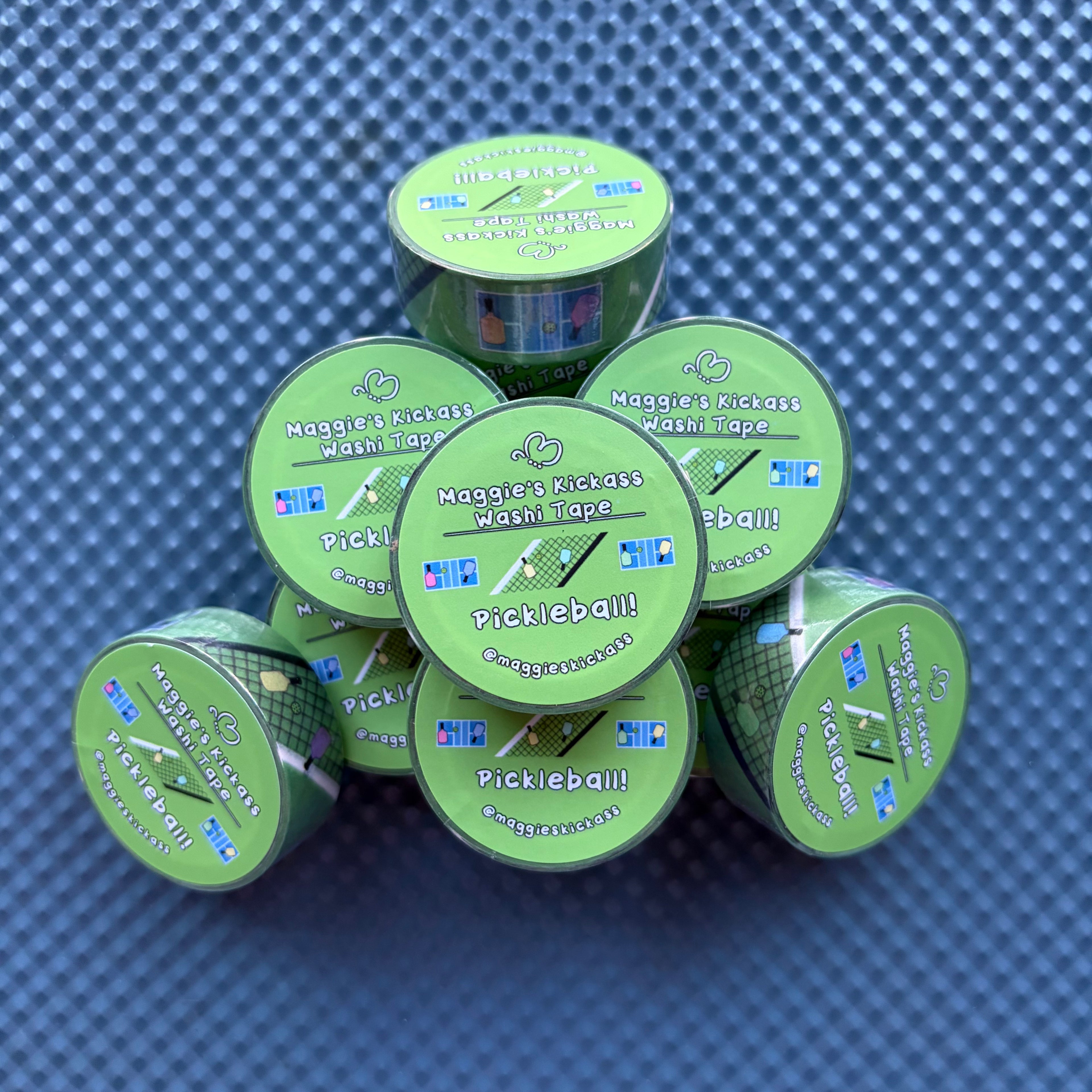 A stack of green washi tape rolls labeled 'Maggie's Kickass Washi Tape' and 'Pickleball!!' on a blue textured background.