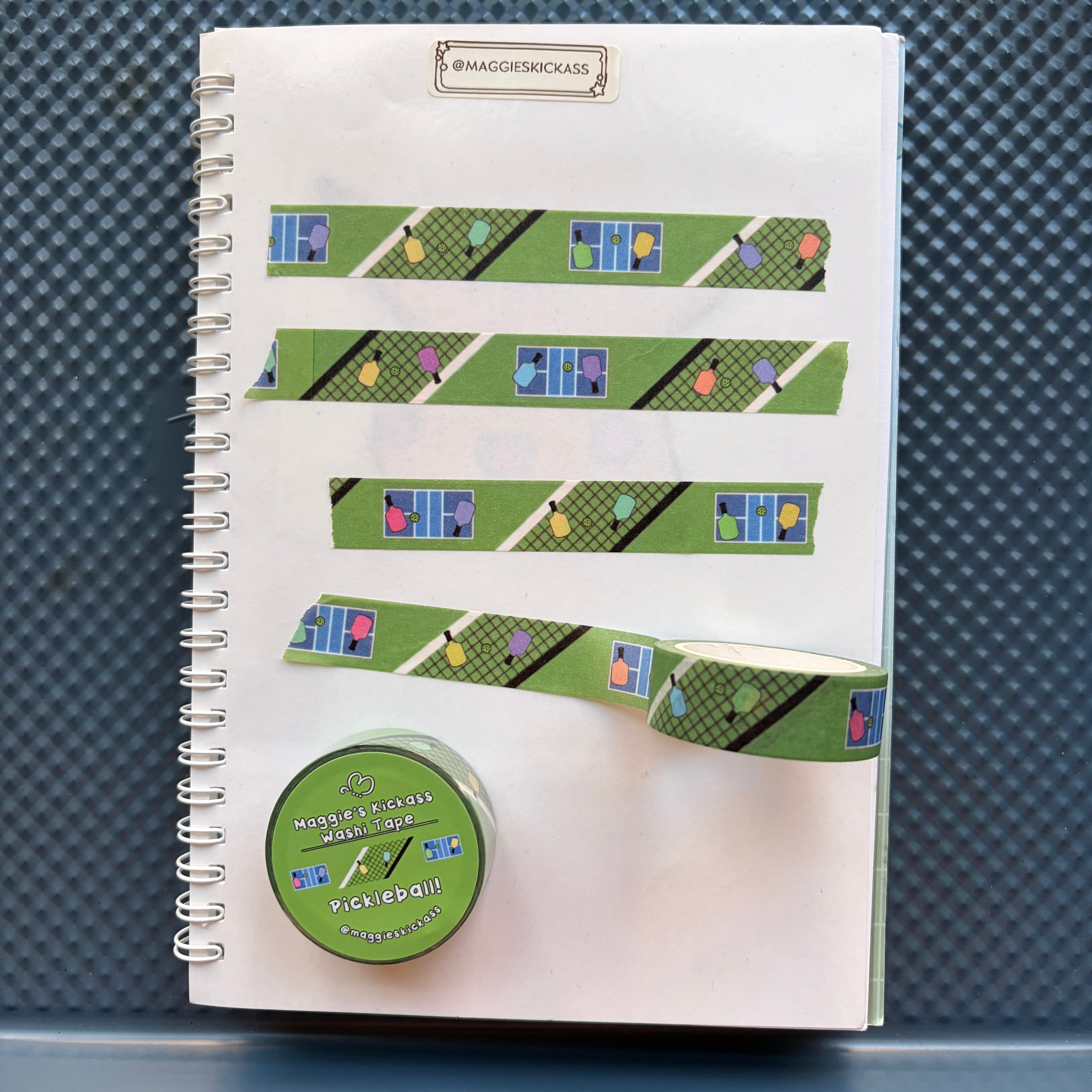 A spiral sticker notebook with green pickleball-themed washi tape strips applied across the pages, plus a roll of Maggie's Kickass Washi Tape Pickleball! washi tape on a dark textured background.